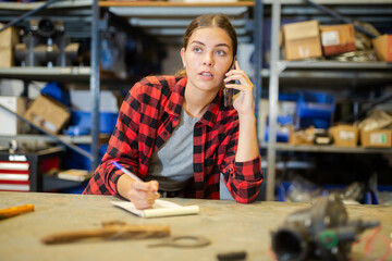 Young woman working in warehouse, making notes while talking on phone.