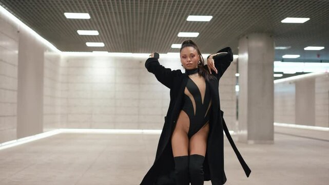 Confident Latina woman fashion model dancing seductively and looking at camera in futuristic design underground subway hall. Female in black bodysuit, thigh-high boots and coat, concrete walls