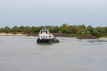 A robust river tugboat navigates a wide, calm river, with tree-lined banks and small islands in the...