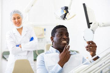 Satisfied patient examines cured teeth using mirror while sitting in a dentist chair