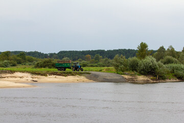 A blue tractor with a large green trailer is parked on a gravel path by a river, with a forest and...