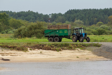 A blue tractor with a large green trailer sits on a gravel road next to a sandy riverbank, with a...