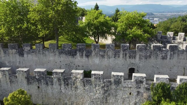 stunning aerial view of Celje Old Castle Celjski Stari Grad perched on a hill, surrounded by lush green forests. The castle overlooks the city of Celje, with the Savinja River winding through, and