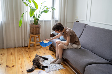 Tired caring woman fan overheating cat on floor in hot apartment with no AC. Pet owner struggle in...