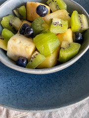 fresh tropical fruit salat desert in blue ceramic bowl on pottery plate and linen napkin, colorful mix pineapple, kiwi, blueberries, high angle view, healthy lifestyle concept