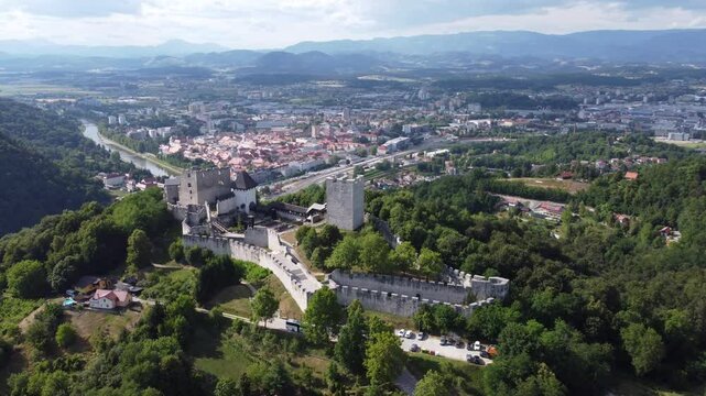 stunning aerial view of Celje Old Castle Celjski Stari Grad perched on a hill, surrounded by lush green forests. The castle overlooks the city of Celje, with the Savinja River winding through, and