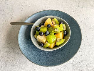 fresh tropical fruit salat dessert in blue ceramic bowl on clay plate on white stone marble background, colorful mix pineapple, kiwi, blueberries, high angle view, copy space