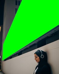 Modern Hijab Woman Listening to Music Under Giant Green Screen Billboard at Night