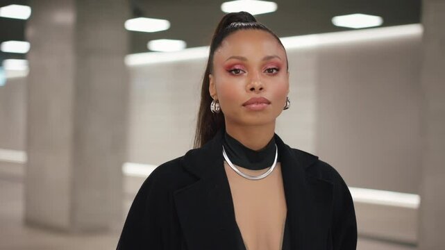 Tracking shot portrait Latina female walking through modern station tunnel hall. Sexy woman wearing long black coat walks seductively looking at camera. Futuristic architecture, concrete walls