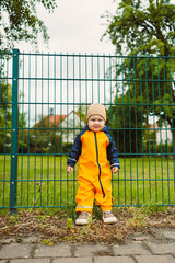 A cheerful and happy child dressed in vibrant rain gear is joyfully standing next to a green fence...