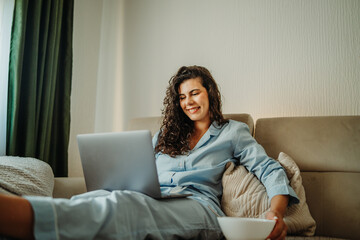 Fototapeta premium Young caucasian woman eating cereals and working on laptop in her pyjamas in the morning 