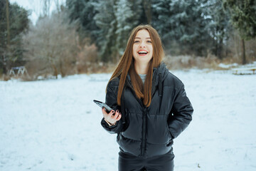 A cheerful young woman is enjoying the beautiful snowy landscape while holding her smartphone,...
