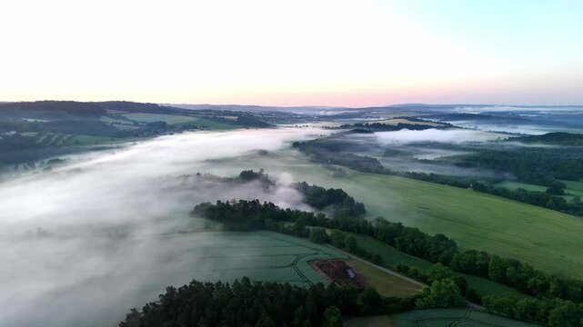 Aerial drone footage of a misty meadow surrounded by forest and mountains at sunrise. Soft golden light filters through fog, creating a tranquil, serene landscape that blends nature&rsquo;s calm with early 
