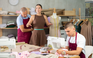 Senior man seamstress employee works with girl client, takes measurements of clients body parts, collects information for creation designer clothing. Guy work with sewing machine in background.