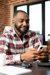 Black male entrepreneur working from home, seated at desk, checking social media on cellphone....