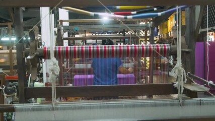 Colorful silk threads on loom with woman weaving in background on handloom in traditional silk factory, Chiang Mai, Thailand. Handmade craftsmanship, artistry of textile production