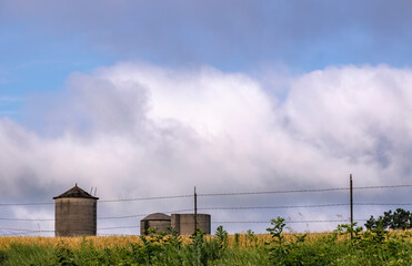 The tops of three concrete silos behind a barbed wire fence with clouds. 