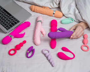 Woman holding sex toy. Various vibrators and laptop on bed. 