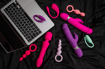 Collection of sex toys on a black sheet. Top view of various vibrators and a laptop. 