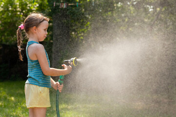 Young girl child spraying water mist with garden hose in sunny backyard on a summer day, copy space...