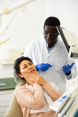 Adult Asian woman suffering from toothache talking to stomatologist while sitting on dental chair...