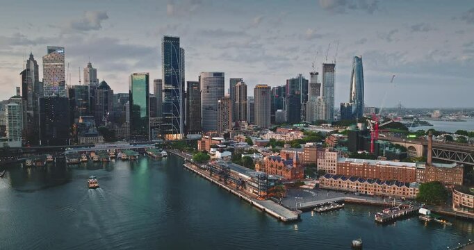 Australia, Sydney: CBD skyline reflecting on calm water in cloudy sunset, modern buildings cityscape skyscrapers towering over ferry cruises sailing ocean harbor. Aerial view drone footage panorama
