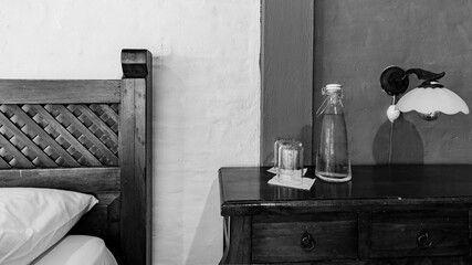 Black and white photo of rustic bedroom with wood bed, nightstand, lamp, glass bottle and glasses....
