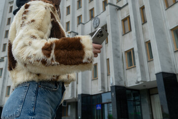 A young individual stands confidently by a contemporary building, wearing a fashionable fur jacket...