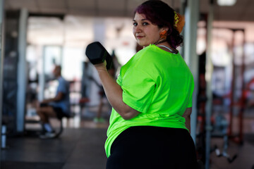 Beautiful overweight Latina woman lifting dumbbells in a gym, wearing a green 80s-style t-shirt,...