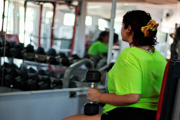 Beautiful overweight Latina woman exercising with dumbbells, looking at herself in the mirror at a...
