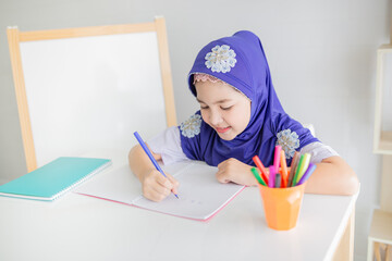 Happy Muslim girl in a purple hijab writing in her notebook at a white desk, with colorful pens beside her. Concept of education, creativity, and childhood development. she doing homework.