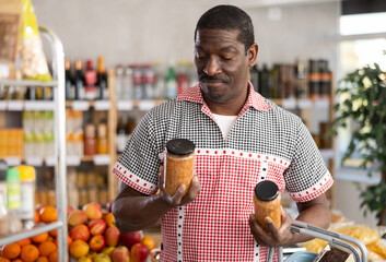African man looking at canned pickled lentils in store. Market visitor choose consider jar with...