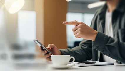 Person using phone at desk with coffee and laptop in office setting.