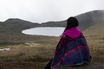 woman wearing a traditional andean clothe sitting on a rock viewing the colombian paramo ecosystem with big colorados lake and mountains at background