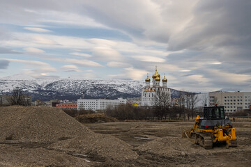Construction work on landscaping a northern city. In the distance, a large cathedral, buildings and snow-covered hills. Clouds in the sky. The city of Magadan, Magadan Region, Far East of Russia.
