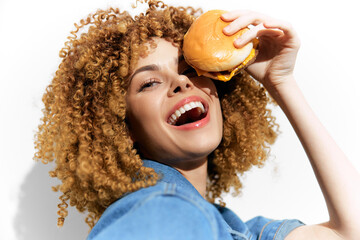 Naklejka premium Happy young woman holding a burger, showcasing joy and excitement in colorful casual wear against a bright background