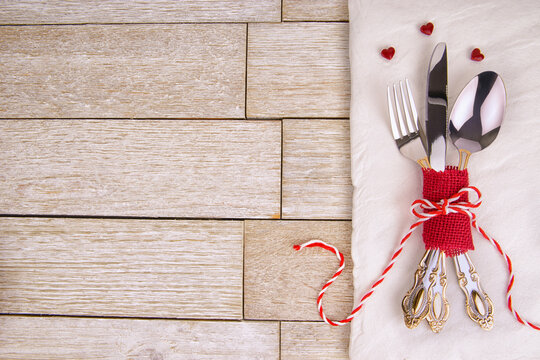 Elegant vintage cutlery tied with red ribbon and tiny hearts on a white napkin over rustic wood. Perfect romantic table setting for Valentine’s Day, wedding dinner or festive love celebration.