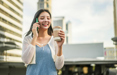 Young Asian successful business woman standing in big city talking on mobile phone and savoring her coffee , Smiling and enjoying a casual conversation, bright daylight, Modern Urban Lifestyle