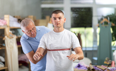 Mature male tailor stands in the workshop and takes measurements of the guy. Client of the atelier...