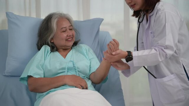 Smiling caring Asian doctor supporting holding hand of olde senior female patient lying on bed at clinic or hospital, Elderly people health care concept