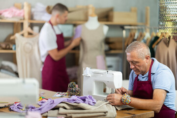 Mature tailor and a young assistant work in a sewing workshop, they sew clothes. Tailor's...