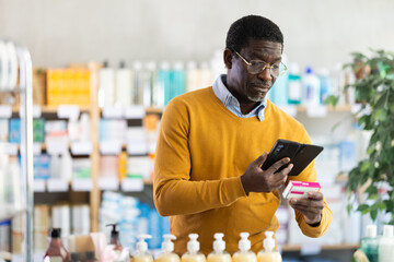 Thoughtful casually dressed middle-aged black man using smartphone to scan barcode on paracetamol box at pharmacy, looking for pain or fever relief information before purchase