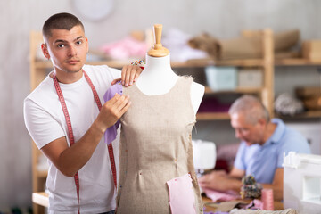 Young guy tailor working with mannequin elderly man assistant sews on machine in workshop