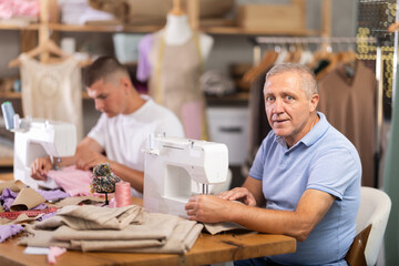 Elderly and young man tailors sewing clothes on sewing machine in workshop