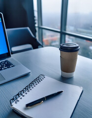 A paper coffee cup placed on a modern office desk with an open laptop, notebook, and pen. Shot from a 45-degree angle. Cool white lighting from large windows. Soft blue and gray tones dominate the sce