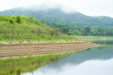 The hillside above the Thai lake reflects a calm summer sky, surrounded by quiet forests and distant mountains.