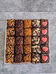 Top view of assorted chocolate brownies in rows, topped with peanuts, choco chips, crumbs, almond slices, and heart-shaped candies. Perfect for dessert, bakery, or festive food themes.

