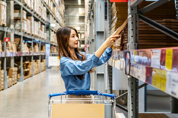 Young asian woman navigates through a vibrant home goods store in warehouse, pushing her shopping cart as she scans the shelves filled with a variety of stylish products, searching for inspiration