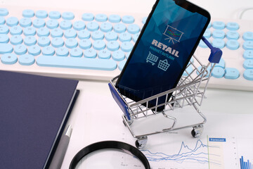 A smartphone displaying Retail Market Outlook report is placed inside a mini trolley, while nearby on the table lies a stack of papers with graphs and charts, accompanied by a book, keyboard and loupe