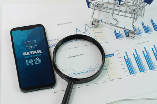A smartphone displaying an Retail Market Outlook report is placed atop a stack of papers with graphs and charts accompanied by a mini trolley and magnifying glass highlighting the report - Powered by Adobe